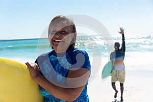 Portrait of smiling african american woman carrying surfboard with senior man waving at beach