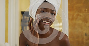 Portrait of smiling african american woman with towel using cream on her face in bathroom