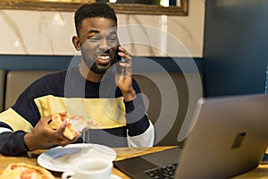 Portrait of smiling african man talking a mobile phone sitting at cafe using laptop and have lunch