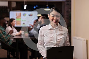 Portrait of smiling asian business employee holding laptop in startup office working overtime