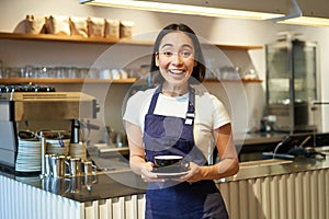 Portrait of smiling asian female barista, making coffee, holding cup of tea and taking it to cafe client, wearing apron