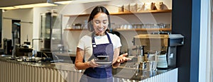 Portrait of smiling asian female barista, making coffee, holding cup of tea and taking it to cafe client, wearing apron