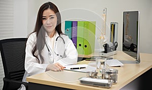 Asian Female Doctor Wearing White Coat With Stethoscope Sitting Behind Desk In Office