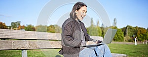 Portrait of smiling asian girl sits on bench in park, talks to friend online via laptop, video chats, using computer to