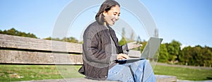 Portrait of smiling asian girl sits on bench in park, talks to friend online via laptop, video chats, using computer to