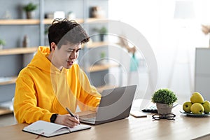 Smiling asian man working on laptop and writing