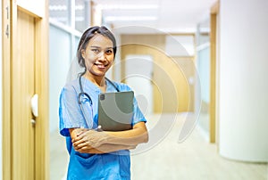 Portrait of smiling asian nurse or female doctor health worker wearing blue uniform holding digital tablet while posing on hallway