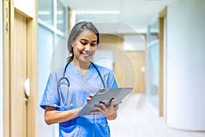 Portrait of smiling asian nurse or female doctor health worker wearing blue uniform using digital tablet while posing on hallway