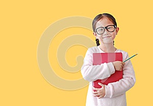 Portrait of smiling Asian schoolgirl wearing eyeglass hug a book and holding pencil in hand isolated over yellow background with