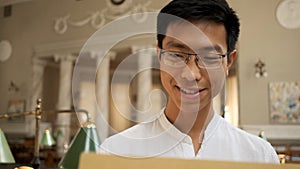 Portrait of smiling asian student happily opening envelope with exam results in university library. Young attractive guy