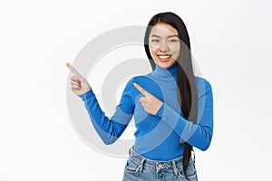 Portrait of smiling asian woman pointing fingers left, showing amazing news, promo banner, offer, standing over white