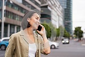 Portrait of smiling asian woman talking on mobile phone, walking on street near busy road and speaking to friend