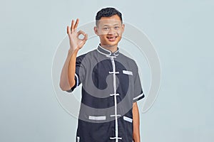 Portrait of smiling Asian young man wearing karate uniform showing okay gesture isolated on gray background