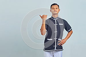 Portrait of smiling Asian young man wearing taekwondo kimono pointing finger at copy space over gray background