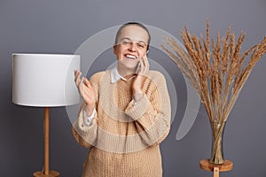 Portrait of smiling attractive positive woman wearing warm sweater standing against gray wall with lamp and dried flower on
