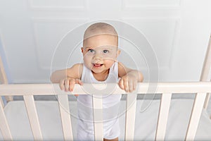 Portrait of a smiling baby boy 8 months old standing in a crib in a children`s room in white clothes and looking at the camera,