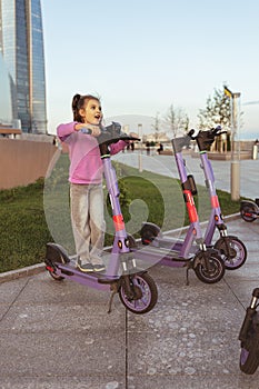 Portrait of smiling baby girl 6-7 years old in pink hoodie on violet scooter, lifestyle, kicks haring