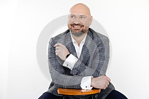 Portrait of smiling bald middle-aged businessman in grey checkered jacket, vest, white shirt, sitting at stool, posing.