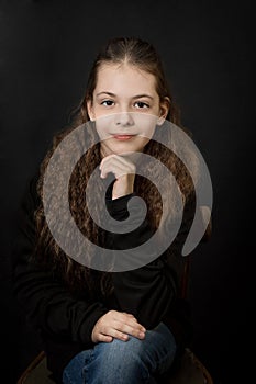 Portrait of a smiling beautiful  brown-haired girl with wavy hair, in black clothes on a black background, sitting on a chair;