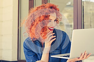 Portrait smiling beautiful city lifestyle woman holding notebook, looking to laptop, touching chin. Pretty young girl red curly