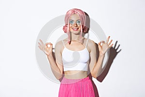 Portrait of smiling beautiful girl with pink anime wig and bright makeup, showing okay signs with satisfied expression