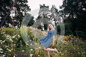 A portrait of a smiling beautiful slender girl with blond long hair holding wildflowers in her hands, in a blue summer dress