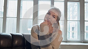 Portrait of smiling beautiful woman looking at camera sitting in armchair opposite large window. A blonde stretches