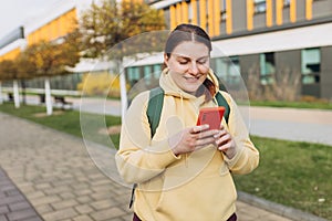 A portrait of a smiling beautiful woman texting sms with her phone on urban background. Happy student with backpack is