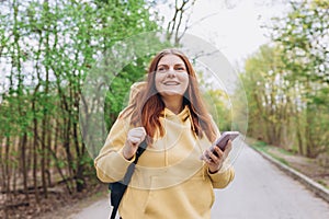 A portrait of a smiling beautiful woman walking with phone on nature background. Happy woman with backpack is using a