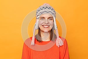 Portrait of smiling beautiful woman wearing orange jumper and knitted earflap hat standing isolated over yellow background,