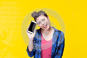 Portrait of a smiling beautiful young asian woman showing blank smartphone screen looking at the camera on yellow background