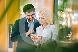 Portrait of smiling beautiful young couple using mobile phone standing in hall of shopping mall.