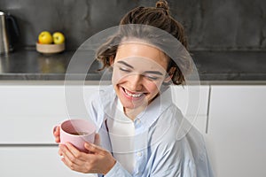 Portrait of smiling, beautiful young woman, drinking coffee in kitchen, morning magic with cuppa tea, looking tenderly