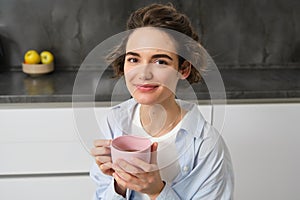 Portrait of smiling, beautiful young woman, drinking coffee in kitchen, morning magic with cuppa tea, looking tenderly