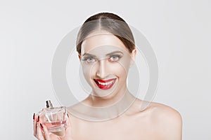 Portrait of smiling beautiful young woman holding showing perfume bottle in her hand, enjoying its smell and looking at camera,