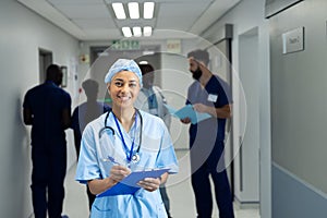 Portrait of smiling biracial female doctor writing notes in busy hospital corridor, copy space
