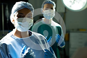 Portrait of smiling biracial female surgeon in cap and mask in operating theatre with colleague