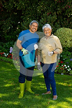 Portrait of smiling biracial senior couple holding watering can and gardening fork standing in yard