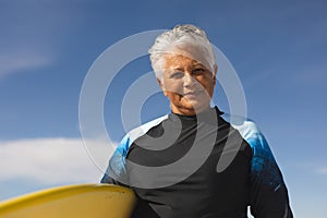 Portrait of smiling biracial senior woman carrying surfboard at beach against blue sky on sunny day