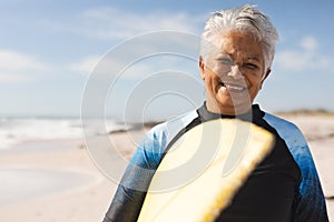Portrait of smiling biracial senior woman carrying surfboard at beach against sky on sunny day