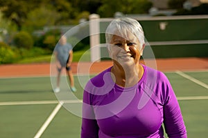 Portrait of smiling biracial senior woman standing in tennis court with senior man in background