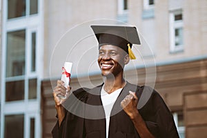 Portrait of smiling black guy graduate from university standing outdoors near building and holding in hand higher