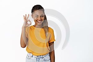 Portrait of smiling Black woman winks and shows okay sign, approves, compliments, recommends company, stands over white