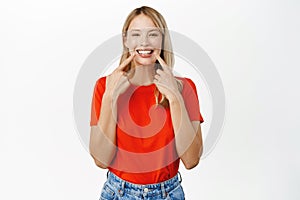 Portrait of smiling blond girl pointing at her white teeth, perfect smile, dental clinic advertisement, standing happy