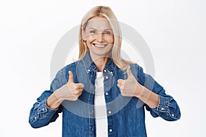 Portrait of smiling blond senior woman, grandmother shows thumbs up, approves smth, praise and compliment, stands over