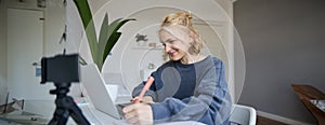 Portrait of smiling blond woman writing in notebook, making notes, recording content for social medial on digital camera
