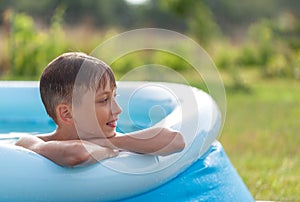 Portrait of smiling boy splashing in an inflatable pool on a sunny summer day in nature. Child staying cool in the summer heat