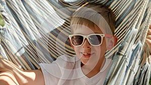 Portrait of smiling boy wearing sunglasses, sways and lounges in a garden hammock, radiating the aura of summertime