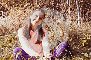 Portrait of a smiling brunette woman sitting in autumnal yellow grass. Hipster girl in fall landscape with dry grass. Cozy autumn