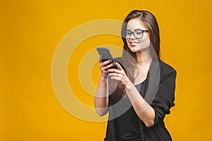 Portrait of smiling business woman in eyeglasses holding smartphone and looking back over yellow background. Using phone